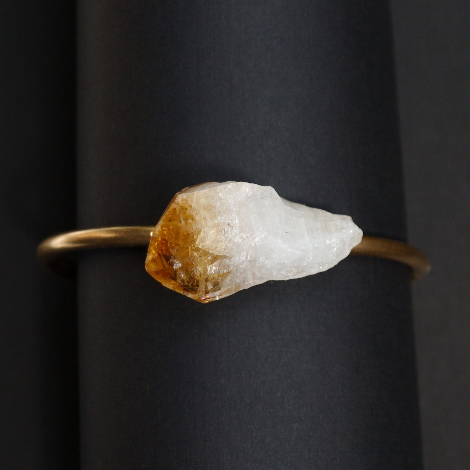 The Raw Citrine Bangle features a stunning large raw crystal centerpiece set against a dark background in an adjustable brass band.