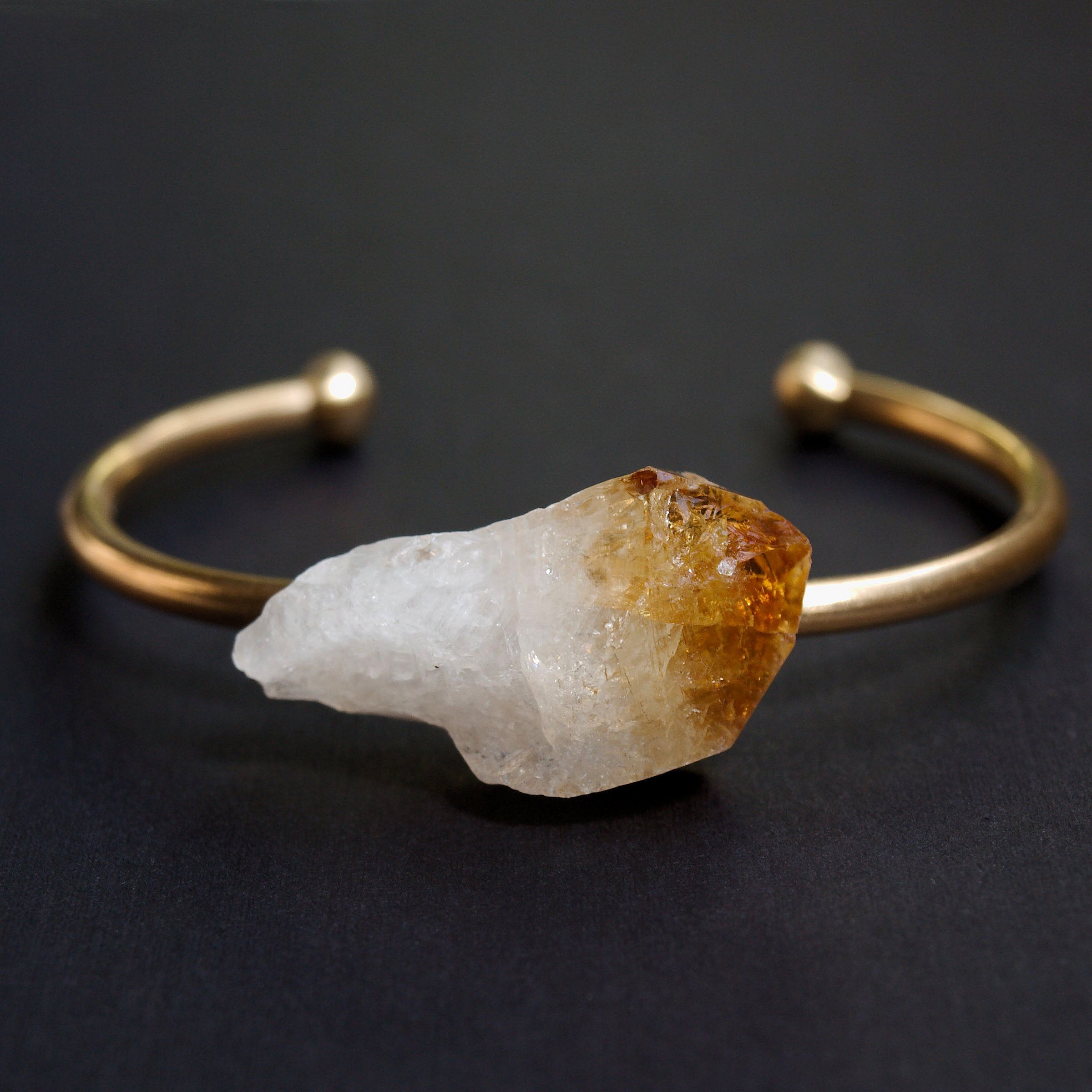 The Raw Citrine Bangle features a stunning large raw crystal centerpiece set against a dark background in an adjustable brass band.