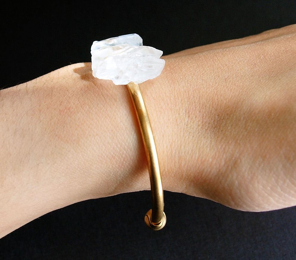The Quartz and Gold Bangle is an open cuff bracelet adorned with five rough white quartz stones on top, symbolizing spiritual growth, set against a black background.