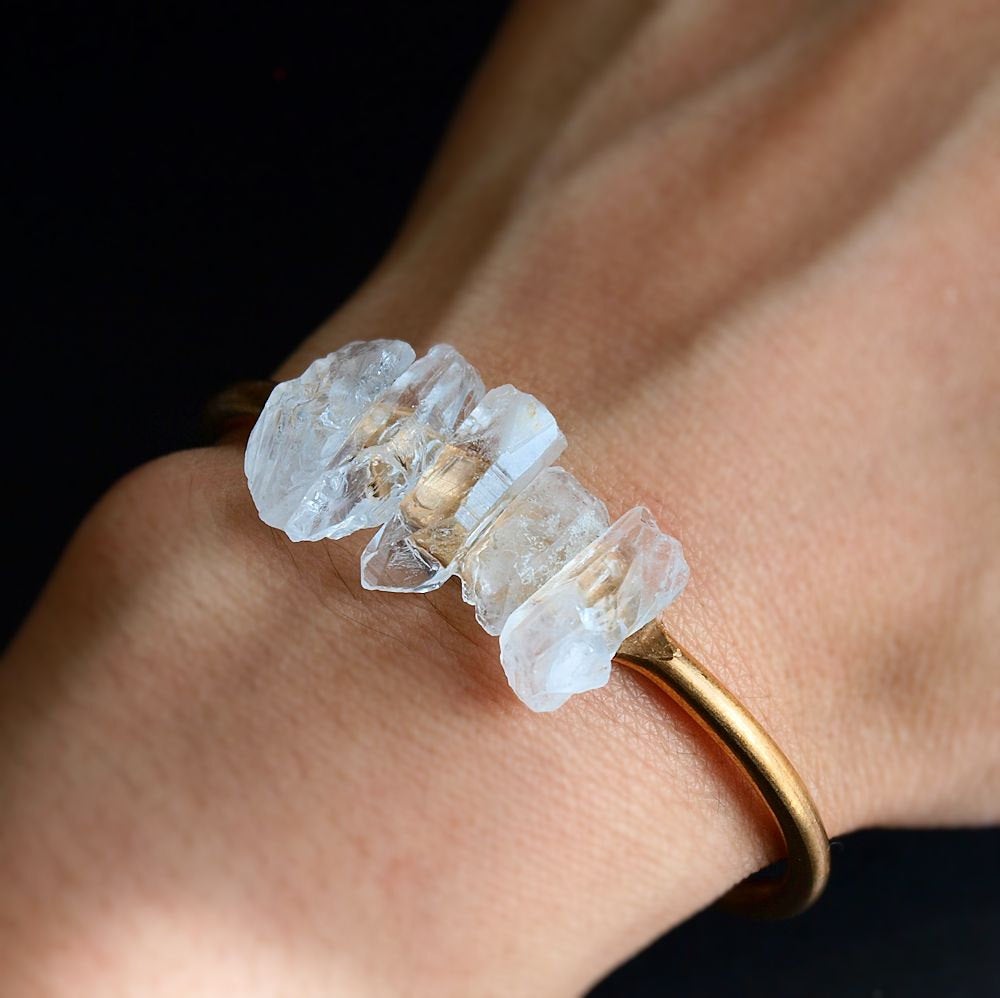 The Quartz and Gold Bangle is an open cuff bracelet adorned with five rough white quartz stones on top, symbolizing spiritual growth, set against a black background.