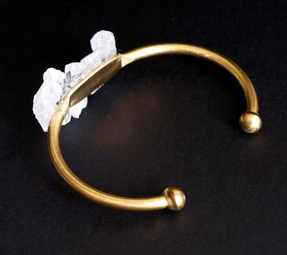 The Quartz and Gold Bangle is an open cuff bracelet adorned with five rough white quartz stones on top, symbolizing spiritual growth, set against a black background.