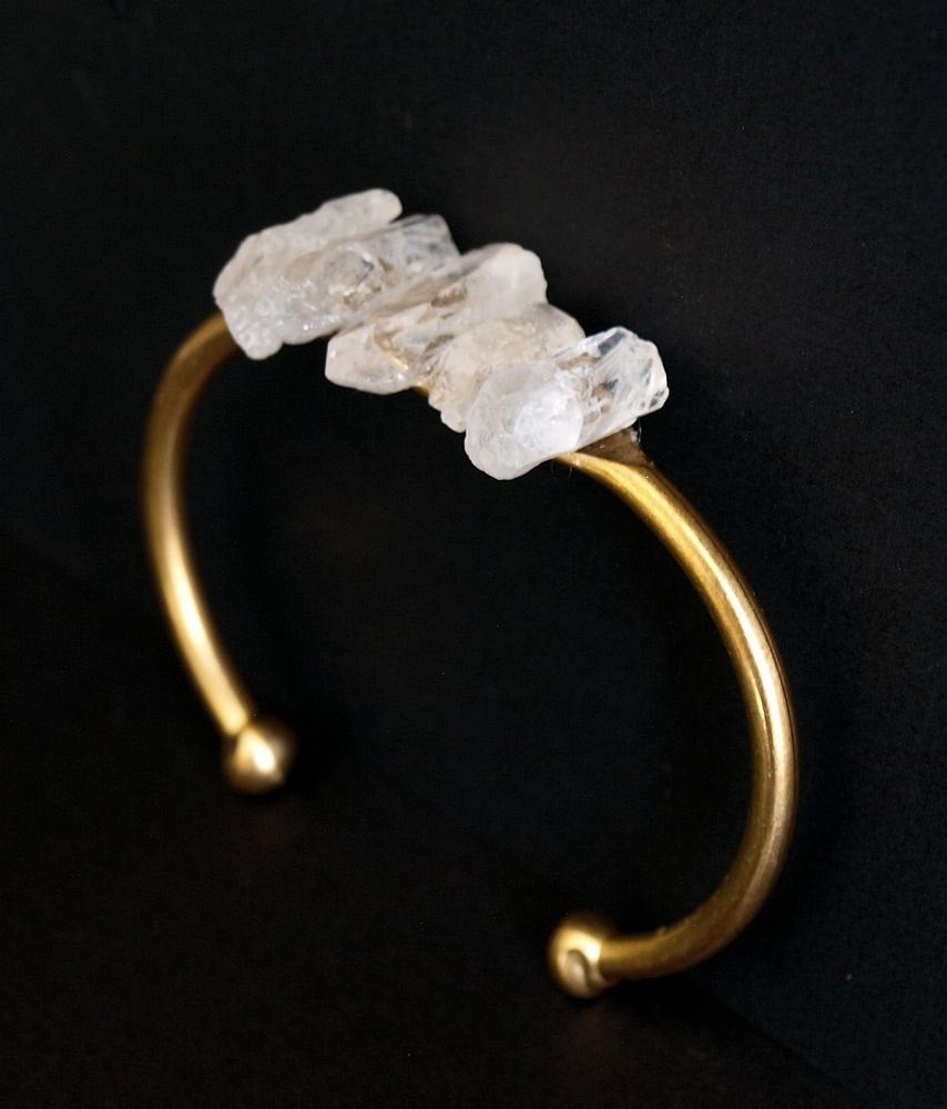 The Quartz and Gold Bangle is an open cuff bracelet adorned with five rough white quartz stones on top, symbolizing spiritual growth, set against a black background.