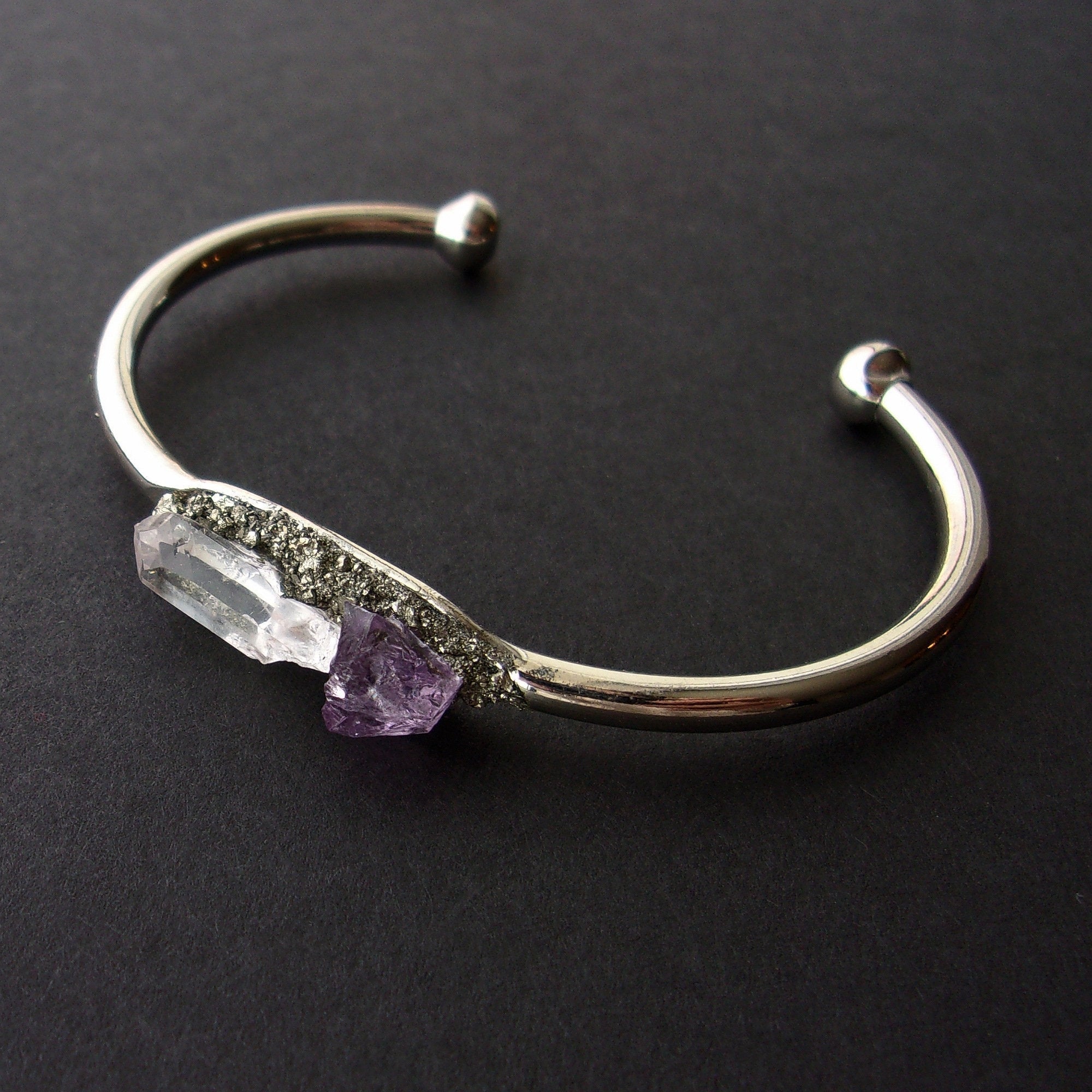 Introducing the Pyrite, Amethyst, and Quartz Bangle—a stunning silver cuff bracelet featuring an asymmetrical design with two raw crystals: a clear quartz famed for its healing properties and a beautiful purple amethyst set against a dark backdrop.