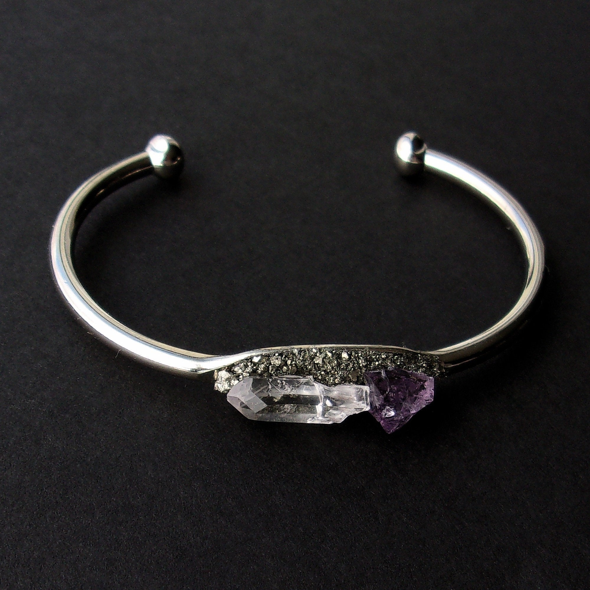 Introducing the Pyrite, Amethyst, and Quartz Bangle—a stunning silver cuff bracelet featuring an asymmetrical design with two raw crystals: a clear quartz famed for its healing properties and a beautiful purple amethyst set against a dark backdrop.
