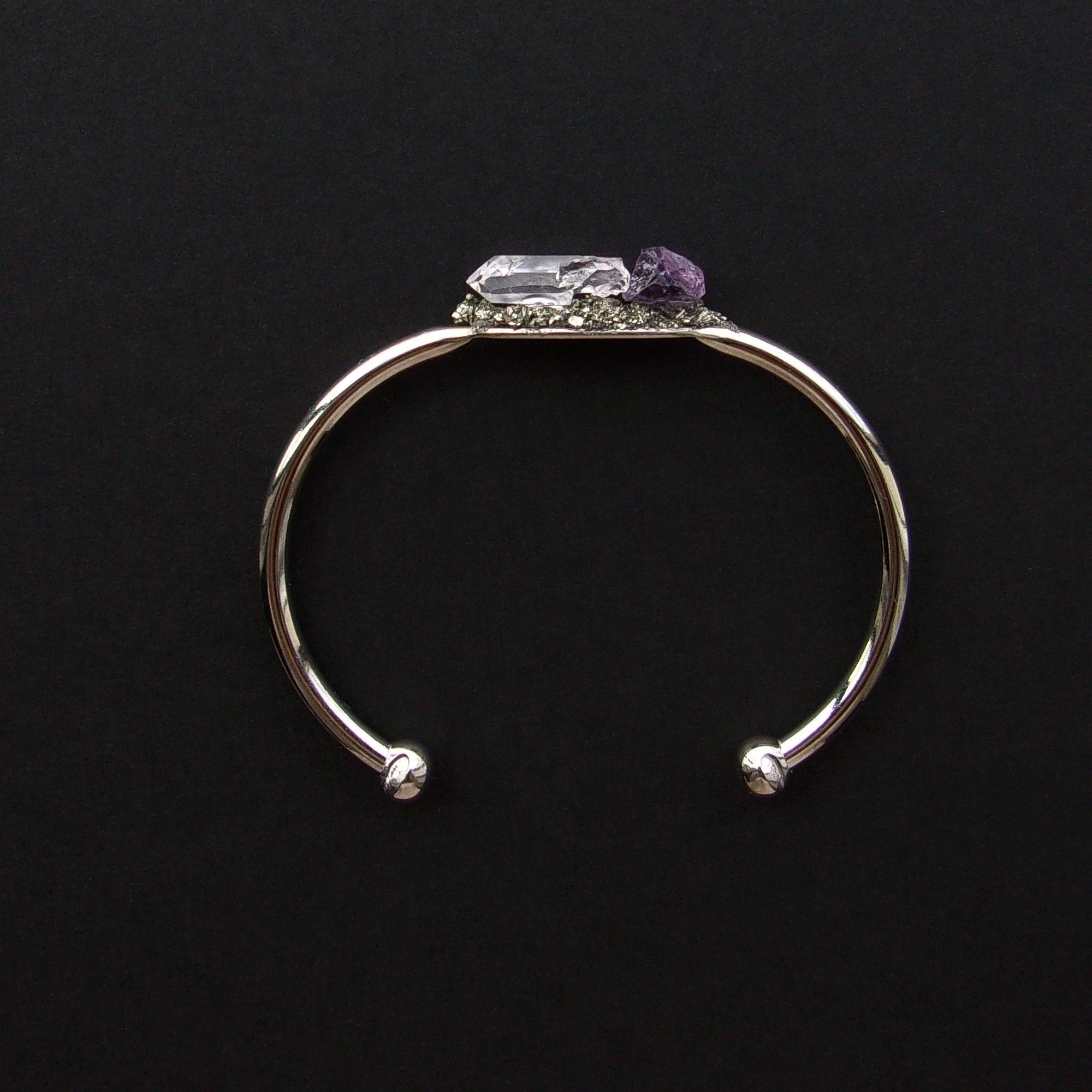 Introducing the Pyrite, Amethyst, and Quartz Bangle—a stunning silver cuff bracelet featuring an asymmetrical design with two raw crystals: a clear quartz famed for its healing properties and a beautiful purple amethyst set against a dark backdrop.