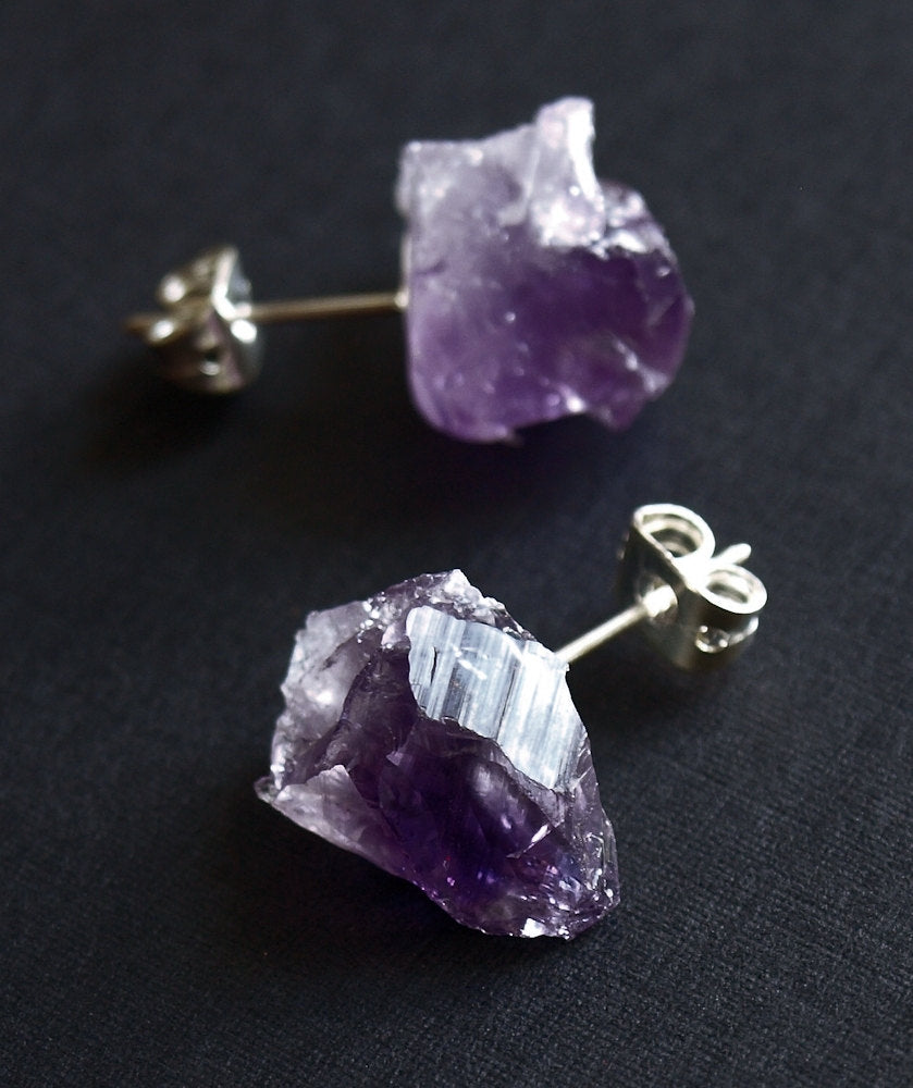 Close-up of the Raw Amethyst Earrings, featuring two raw stud amethysts with surgical steel posts, set against a dark background.