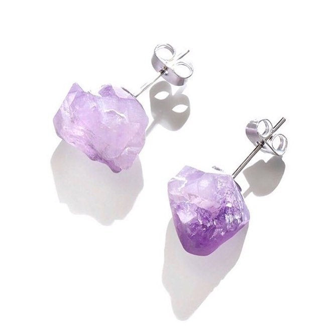 Close-up of the Raw Amethyst Earrings, featuring two raw stud amethysts with surgical steel posts, set against a dark background.