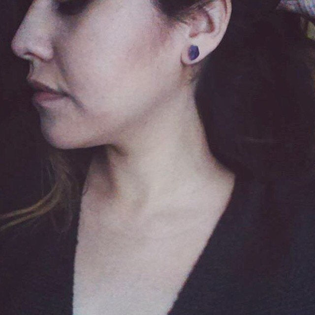 Close-up of the Raw Amethyst Earrings, featuring two raw stud amethysts with surgical steel posts, set against a dark background.