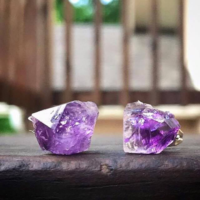 Close-up of the Raw Amethyst Earrings, featuring two raw stud amethysts with surgical steel posts, set against a dark background.