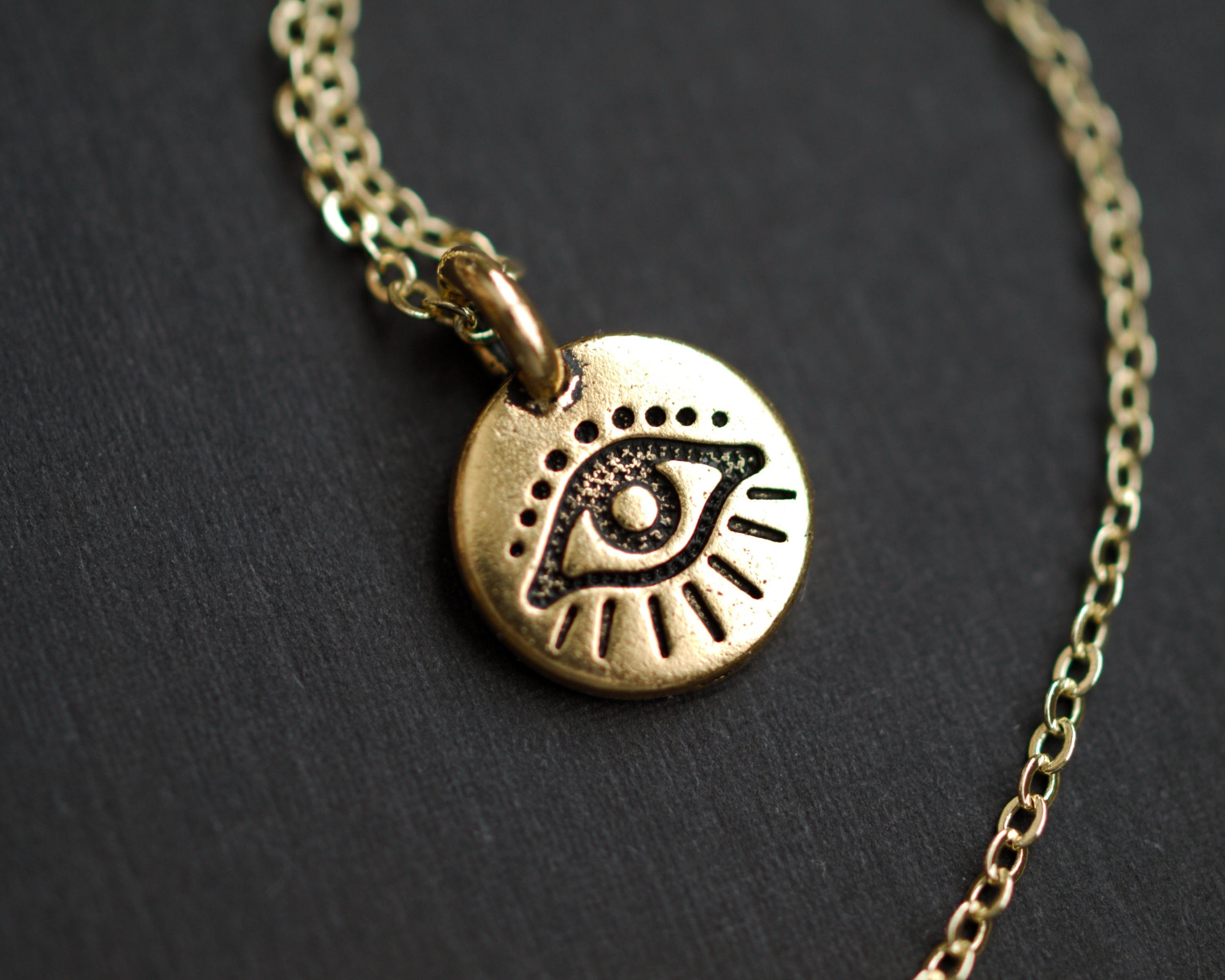 The Tiny Evil Eye Necklace features an eye-shaped pendant on a gold chain, creating a striking contrast against the dark surface, making it a perfect addition to your style.
