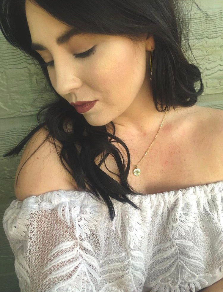The Tiny Evil Eye Necklace features an eye-shaped pendant on a gold chain, creating a striking contrast against the dark surface, making it a perfect addition to your style.
