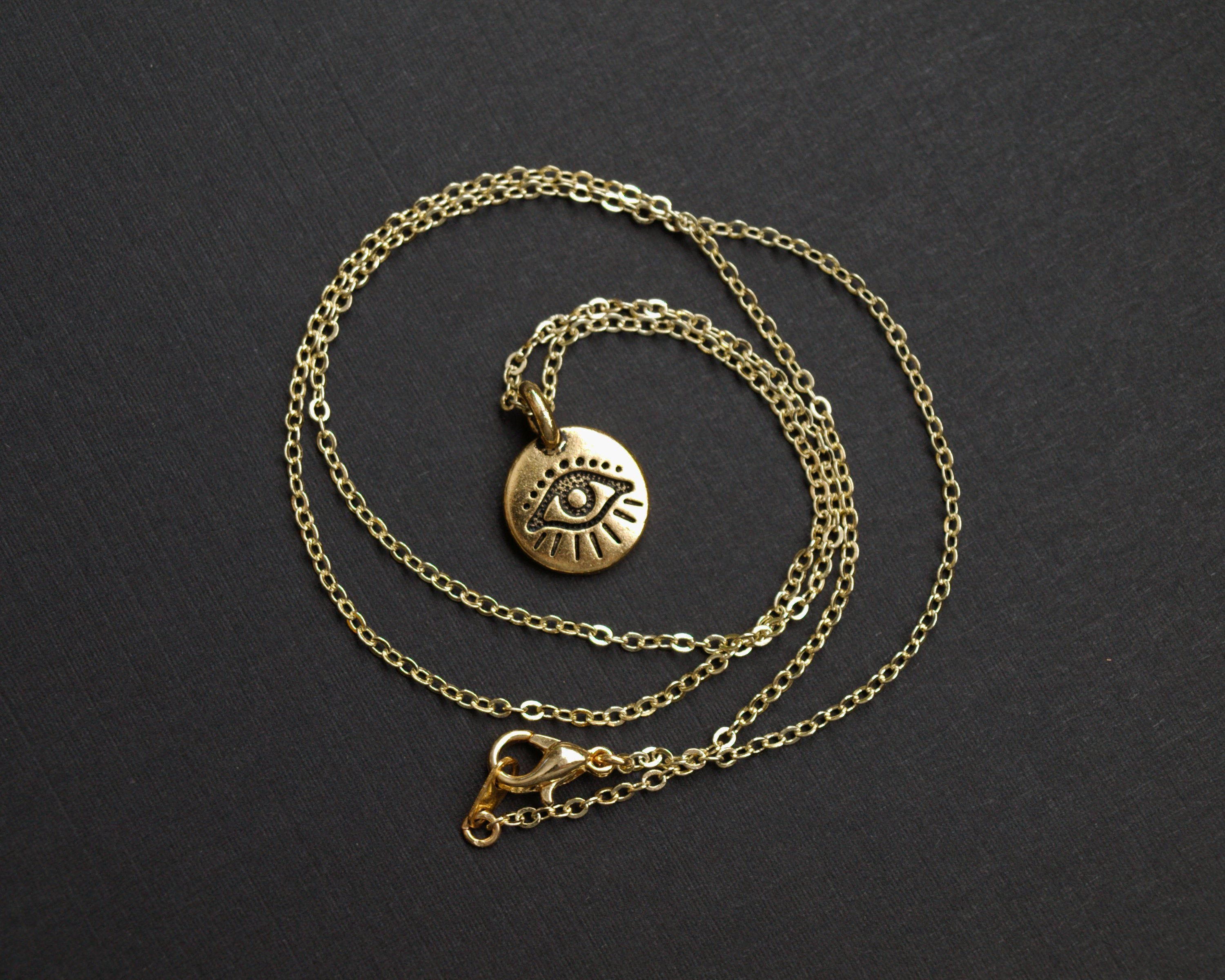The Tiny Evil Eye Necklace features an eye-shaped pendant on a gold chain, creating a striking contrast against the dark surface, making it a perfect addition to your style.