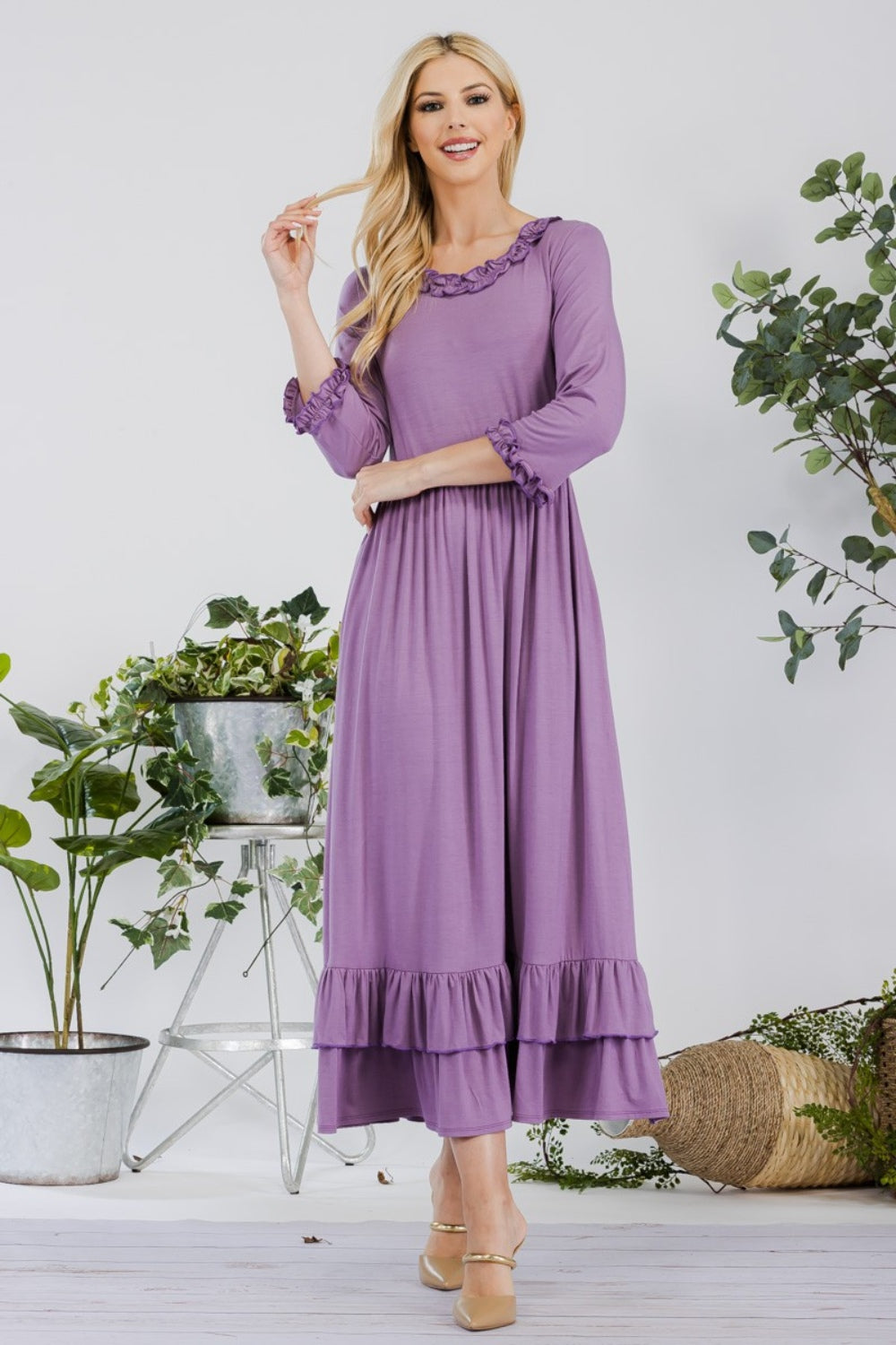 A woman in the Celeste Full Size Layered Ruffle Hem Dress with Pockets, featuring an A-line silhouette and 3/4 sleeves, stands on a wooden floor surrounded by potted plants and a wicker basket.