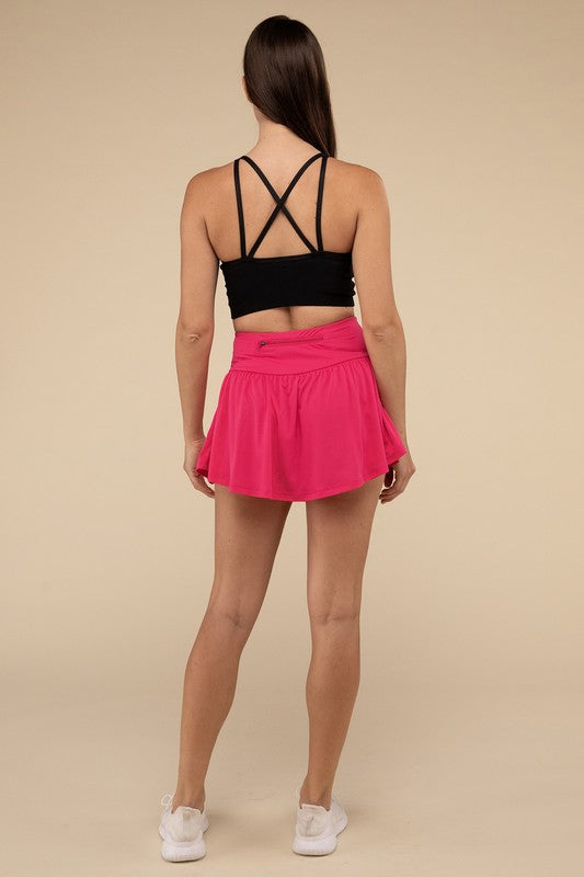 Close-up of a woman in a black crop top and Wide Band Tennis Skirt with Zippered Back Pocket, pulling the skirt to show matching shorts with a zippered back pocket underneath.