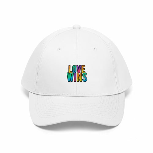 A bearded man wears the Rainbow Love Wins Unisex Twill Cap adorned with the phrase "Rainbow Love Wins" in vibrant colors, along with a gray sweatshirt, as he gazes into the distance against a simple backdrop.