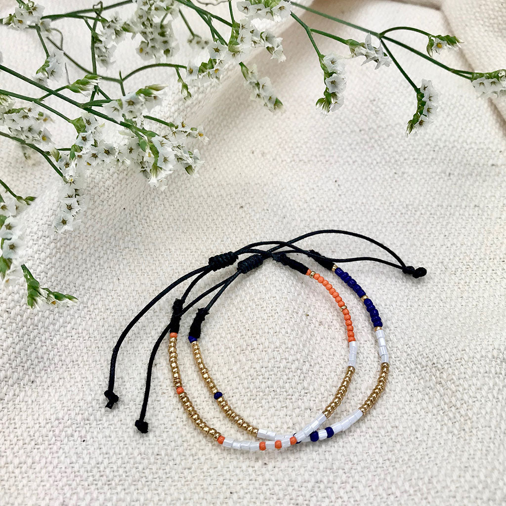 The Dori Beaded Bracelets, one showcasing orange, white, and gold beads and the other blue, white, and gold beads with adjustable cords, are elegantly displayed on a white background. These handmade jewels bring a sophisticated flair to any outfit.