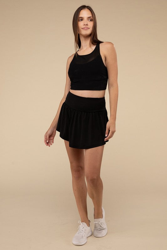Close-up of a woman in a black crop top and Wide Band Tennis Skirt with Zippered Back Pocket, pulling the skirt to show matching shorts with a zippered back pocket underneath.
