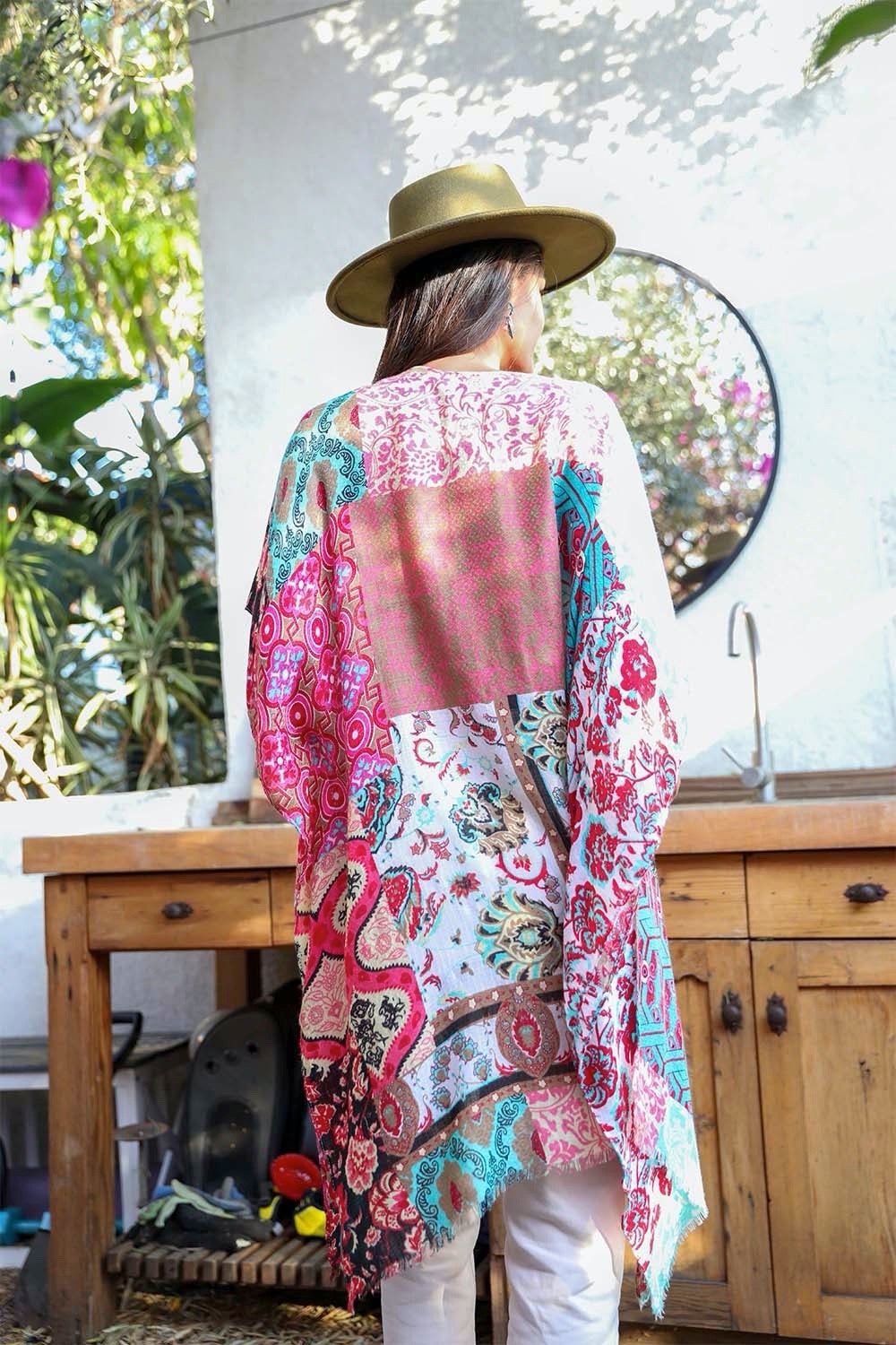 A person in a Boho Patchwork Kimono over their everyday outfit and a hat stands in an outdoor area with a wooden sink and mirror.