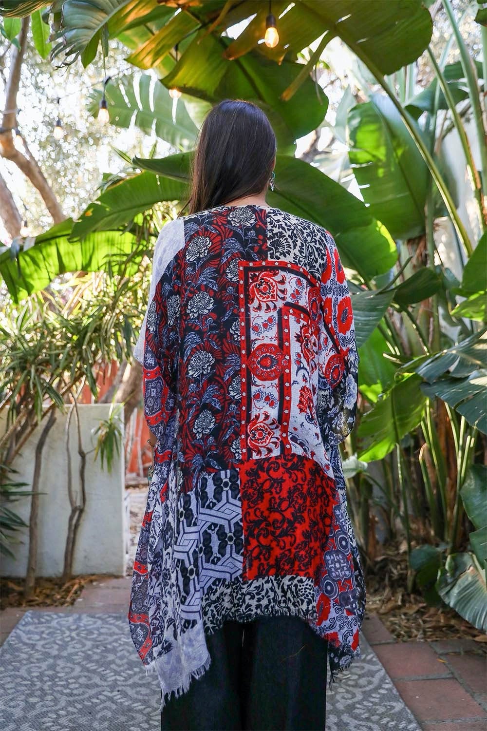 A person in a Boho Patchwork Kimono over their everyday outfit and a hat stands in an outdoor area with a wooden sink and mirror.