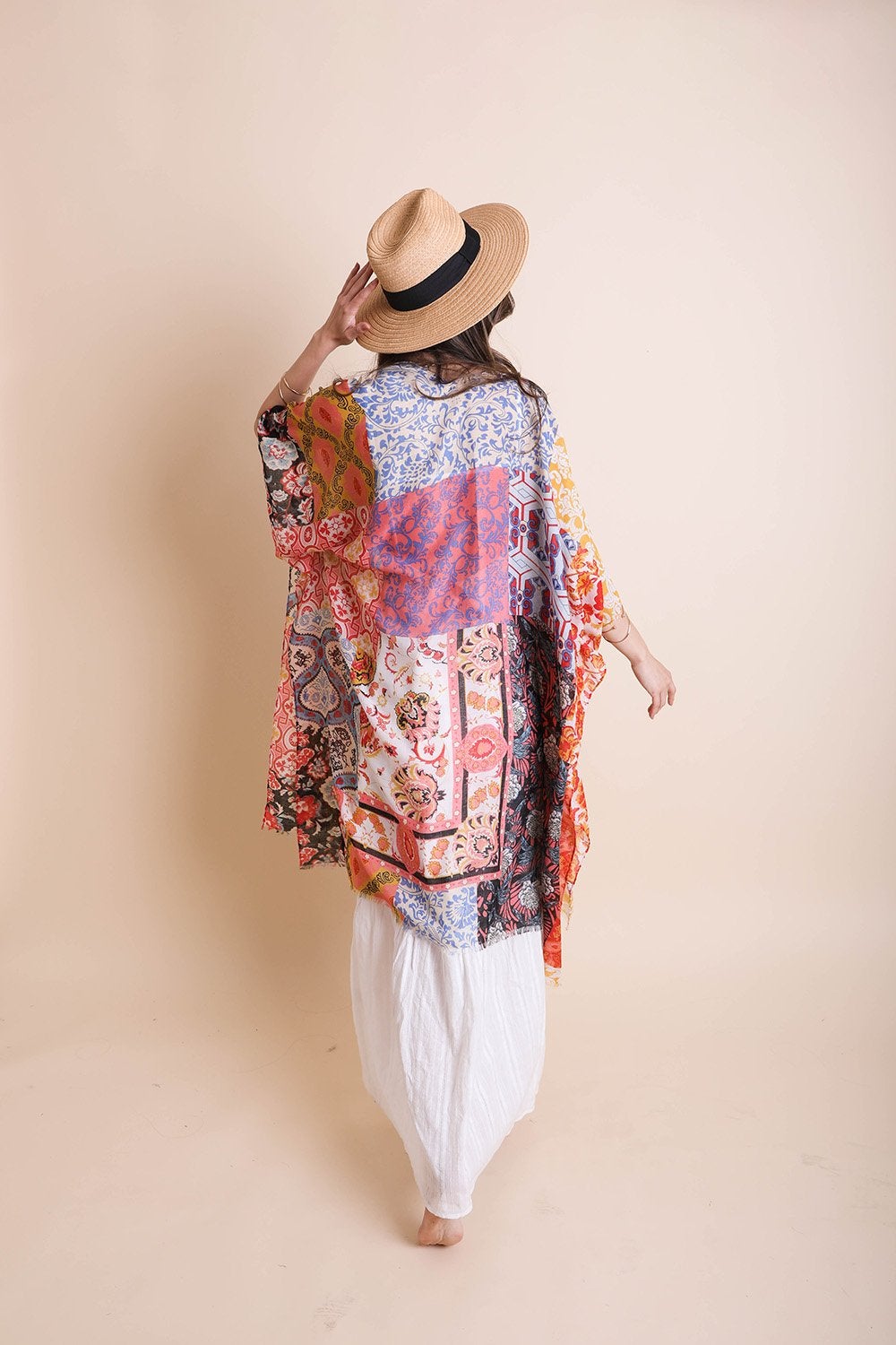 A person in a Boho Patchwork Kimono over their everyday outfit and a hat stands in an outdoor area with a wooden sink and mirror.