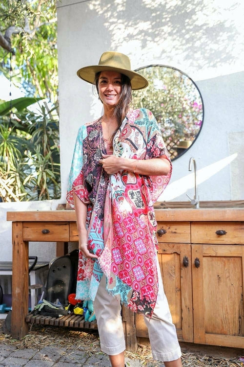 A person in a Boho Patchwork Kimono over their everyday outfit and a hat stands in an outdoor area with a wooden sink and mirror.