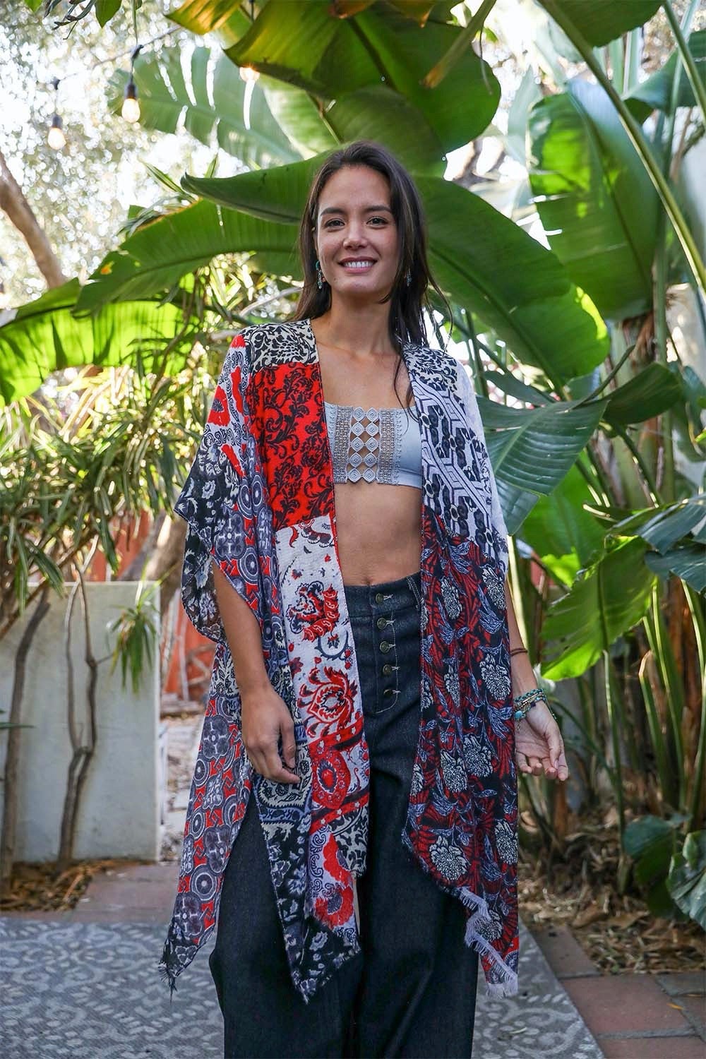 A person in a Boho Patchwork Kimono over their everyday outfit and a hat stands in an outdoor area with a wooden sink and mirror.