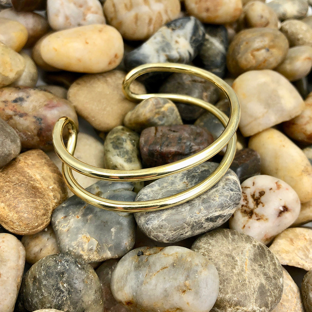 The Bombshell Wire Cuff is a gold double band cuff bracelet with an open-ended design, meticulously handmade in Cambodia. Crafted from recycled brass jewelry materials, this adjustable cuff combines both style and sustainability.