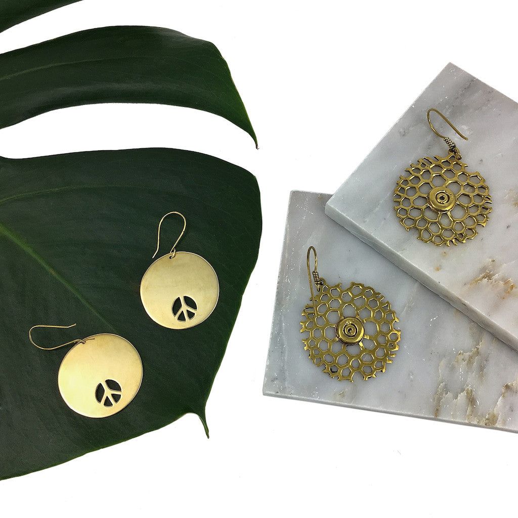 Crafted in Cambodia from recycled brass bombshells, the Bombshell Peace Earrings feature a gold-colored circular peace sign design with cutouts and hook attachments.