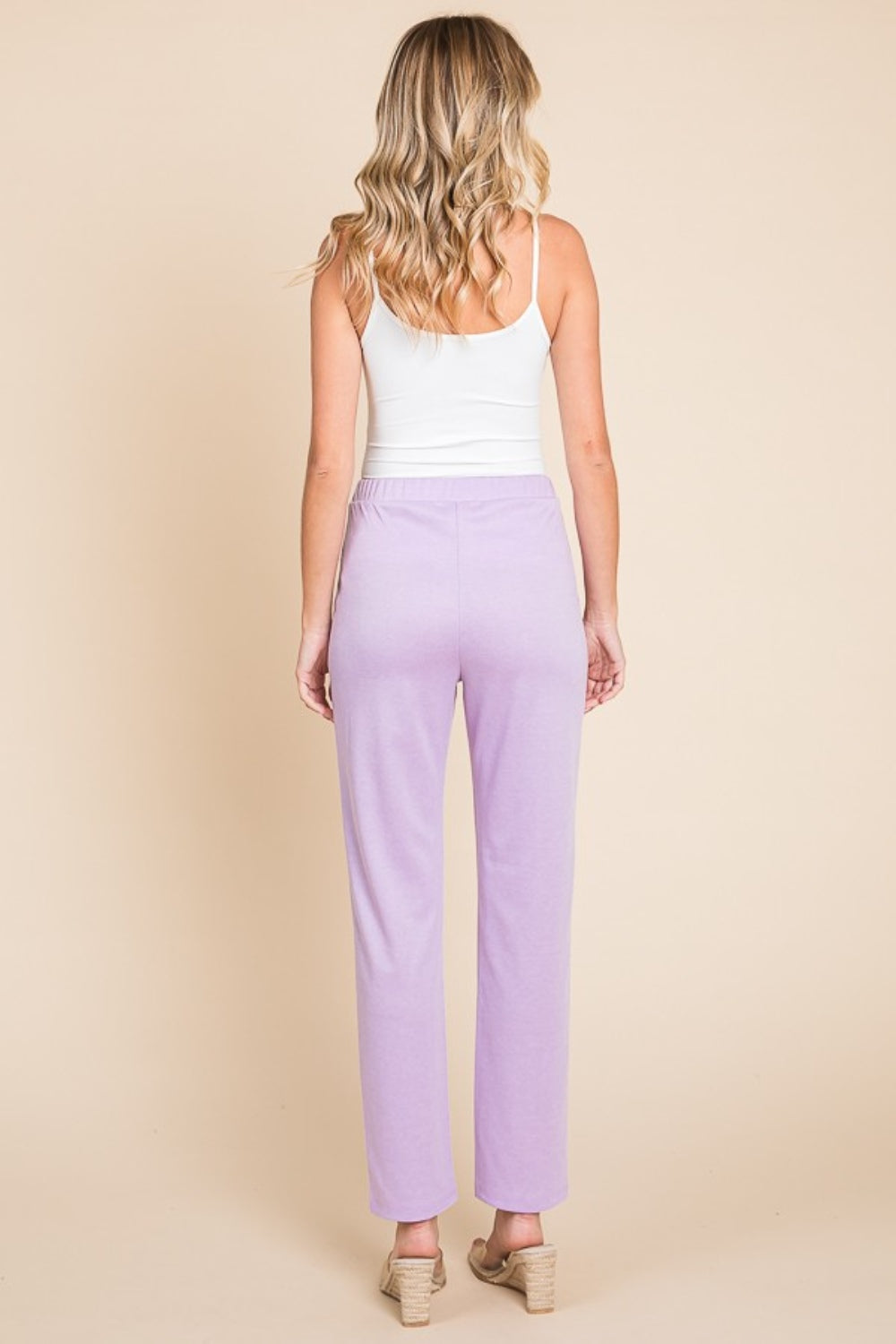 A person wearing a white tank top and the stylish Culture Code Full Size Pin Tuck Detail Slim Pants in a versatile lavender shade stands against a beige background, smiling with hands in pockets.