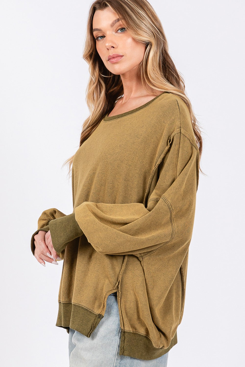 A person wearing a loose, oversized mineral wash SAGE + FIG Side Slit Sweatshirt in olive green, light blue jeans, and a black cap poses against a plain background.