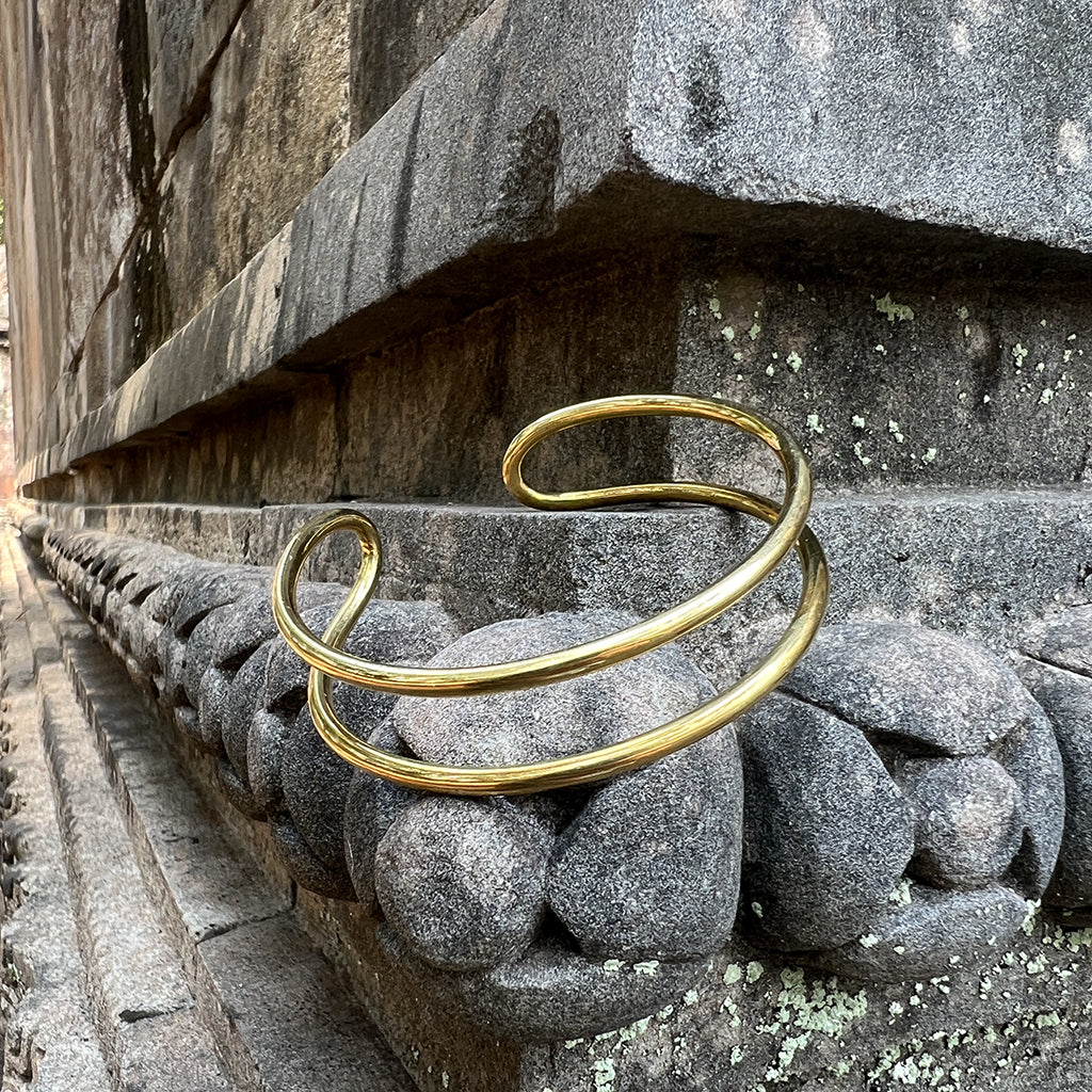 The Bombshell Wire Cuff is a gold double band cuff bracelet with an open-ended design, meticulously handmade in Cambodia. Crafted from recycled brass jewelry materials, this adjustable cuff combines both style and sustainability.