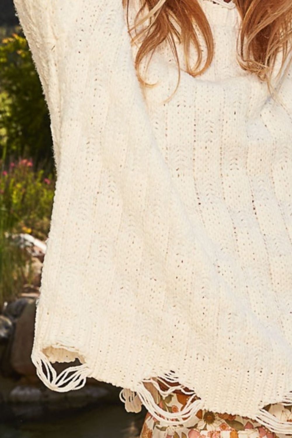 A person with long blonde hair is wearing a white POL Checkered Distressed Edge Sweater and patterned pants, smiling with arms raised outdoors. The slightly oversized fit of the sweater adds a touch of relaxed elegance to her look.