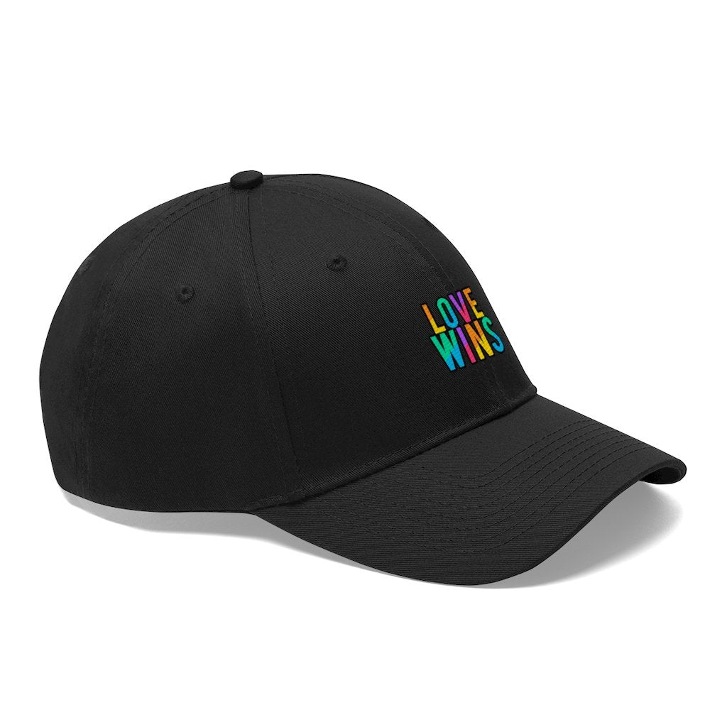 A bearded man wears the Rainbow Love Wins Unisex Twill Cap adorned with the phrase "Rainbow Love Wins" in vibrant colors, along with a gray sweatshirt, as he gazes into the distance against a simple backdrop.