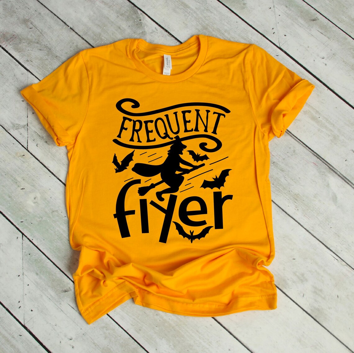 Frequent Flyer Halloween T-shirt in yellow, displayed on a white wooden floor, showcasing a black graphic of a witch flying on a broomstick with bats and the text "Frequent Flyer." This shirt features premium quality soft textile flex print for ultimate comfort.