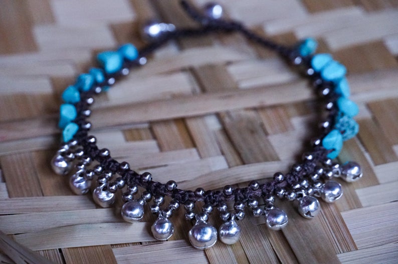 A woman's feet adorned with a Turquoise Silver Bell Boho Anklet are standing on rocks next to a body of water.