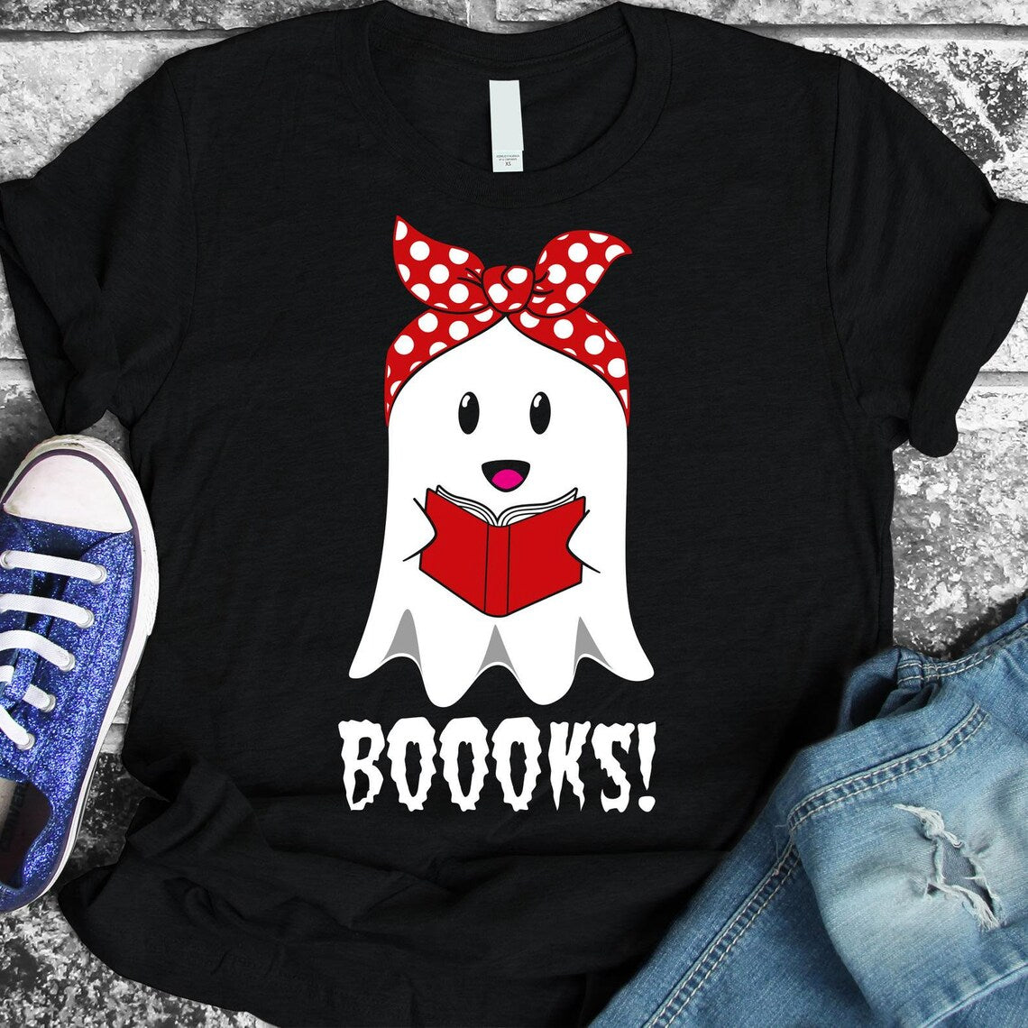 The Boooks Halloween T-shirt is a premium quality, black t-shirt featuring a cartoon ghost with a red bow, holding a red book, and the word "BOOOKS!" underneath. Accompanying the t-shirt are blue glitter sneakers and blue jeans, both offering soft textile flex print for enhanced comfort.