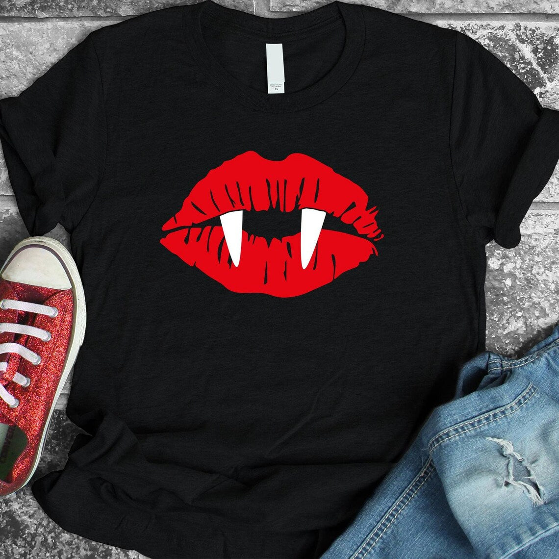 The Vampire Lips Halloween T-shirt in black showcases a striking graphic of red lips with white vampire fangs. The high-quality print is vibrant and pairs perfectly with red sneakers and blue jeans. Crafted from ring-spun cotton, it ensures ultimate comfort.