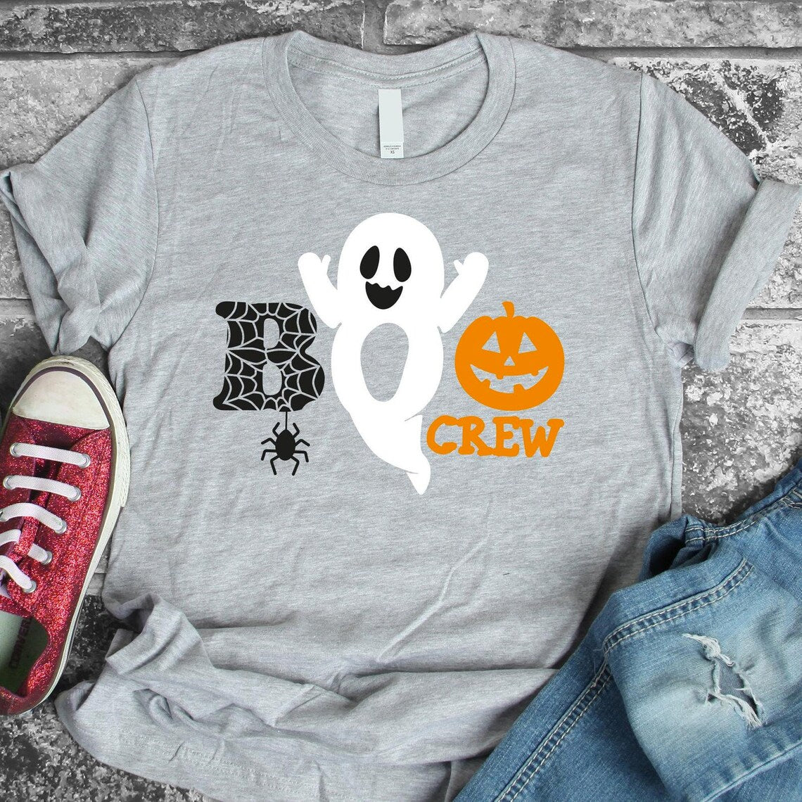 The Boo Crew T-shirt is a premium quality gray tee featuring a Halloween design with a ghost, spider web, and jack-o'-lantern, along with the text "BOO CREW." This Bella Canvas shirt pairs perfectly with red sneakers and blue jeans.