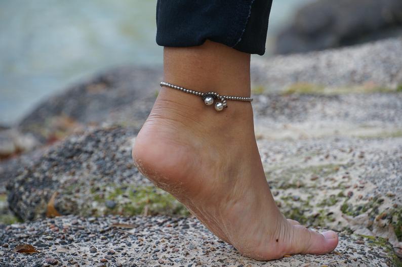 A woman's feet adorned with a Turquoise Silver Bell Boho Anklet are standing on rocks next to a body of water.