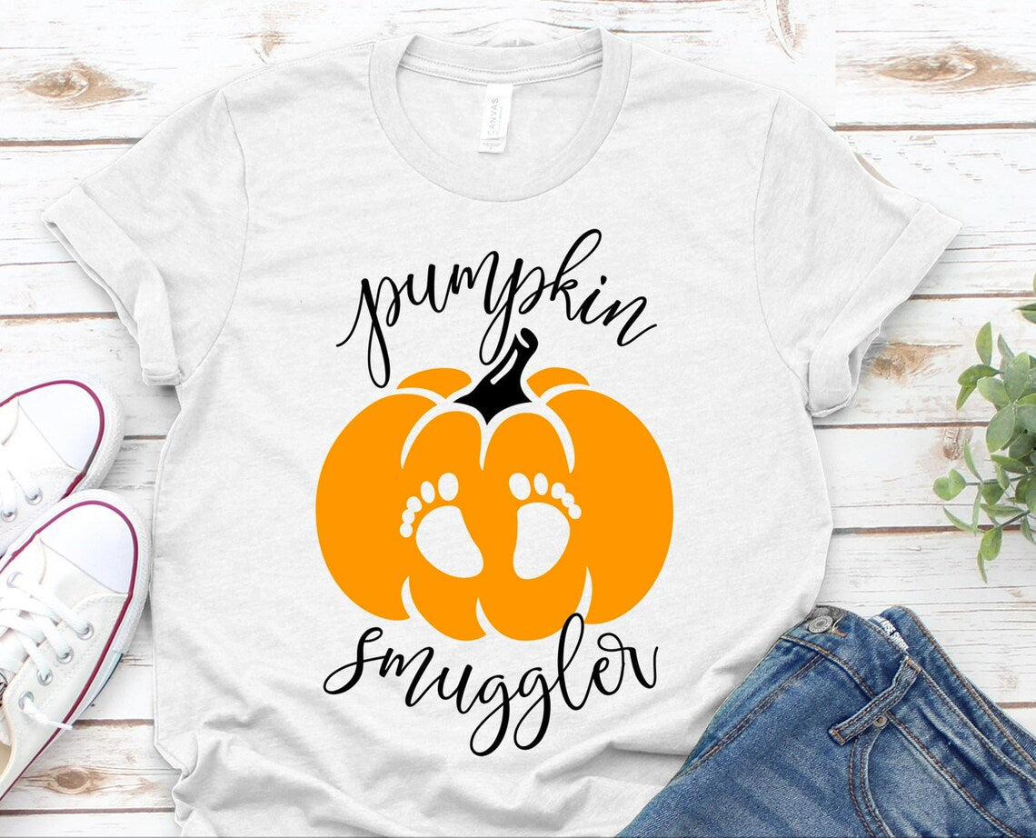 The Pumpkin Smuggler Halloween T-shirt, made from premium quality cotton, features a graphic of an orange pumpkin and baby footprints along with the caption "pumpkin smuggler." It is laid out on a wooden surface beside white sneakers and blue jeans, with a plant in the corner. Enjoy its comfortable retail fit and soft textile flex print.