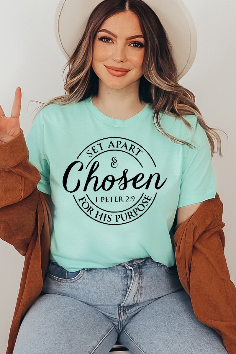 A woman wearing the Chosen T-shirt, featuring the text "Set Apart & Chosen - 1 Peter 2:9 For His Purpose" in mint green premium quality fabric, poses with a peace sign. She has a brown jacket over her shoulders, wavy hair, and a hat. She radiates comfort in this soft textile flex print design.