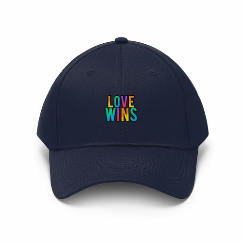 A bearded man wears the Rainbow Love Wins Unisex Twill Cap adorned with the phrase "Rainbow Love Wins" in vibrant colors, along with a gray sweatshirt, as he gazes into the distance against a simple backdrop.
