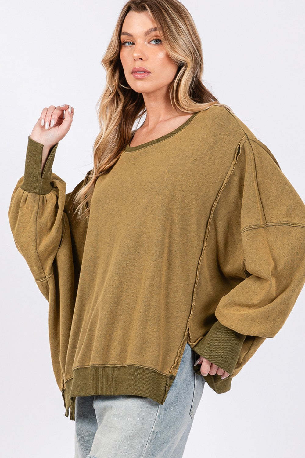 A person wearing a loose, oversized mineral wash SAGE + FIG Side Slit Sweatshirt in olive green, light blue jeans, and a black cap poses against a plain background.