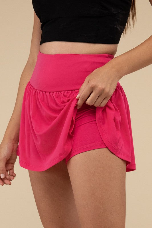 Close-up of a woman in a black crop top and Wide Band Tennis Skirt with Zippered Back Pocket, pulling the skirt to show matching shorts with a zippered back pocket underneath.