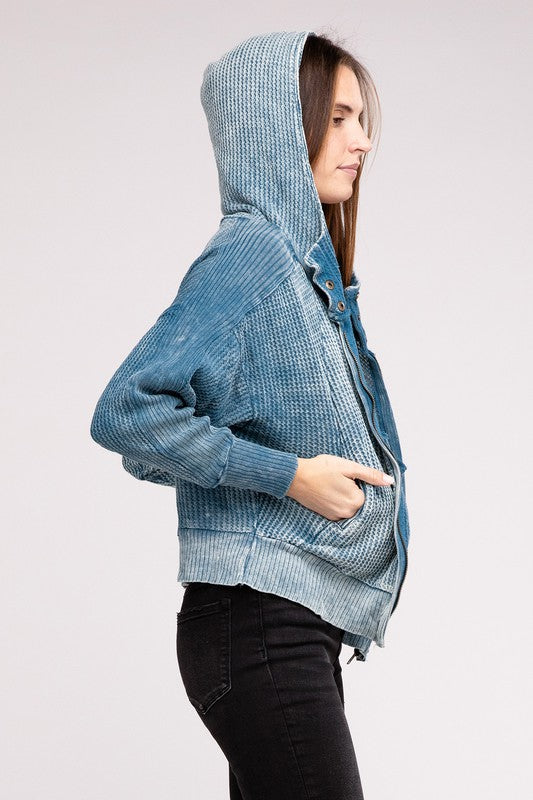 Woman in a Acid Wash Cotton Waffle Hooded Zip-Up Jacket with pockets and black pants, smiling and posing with hands on hips, against a light gray background.