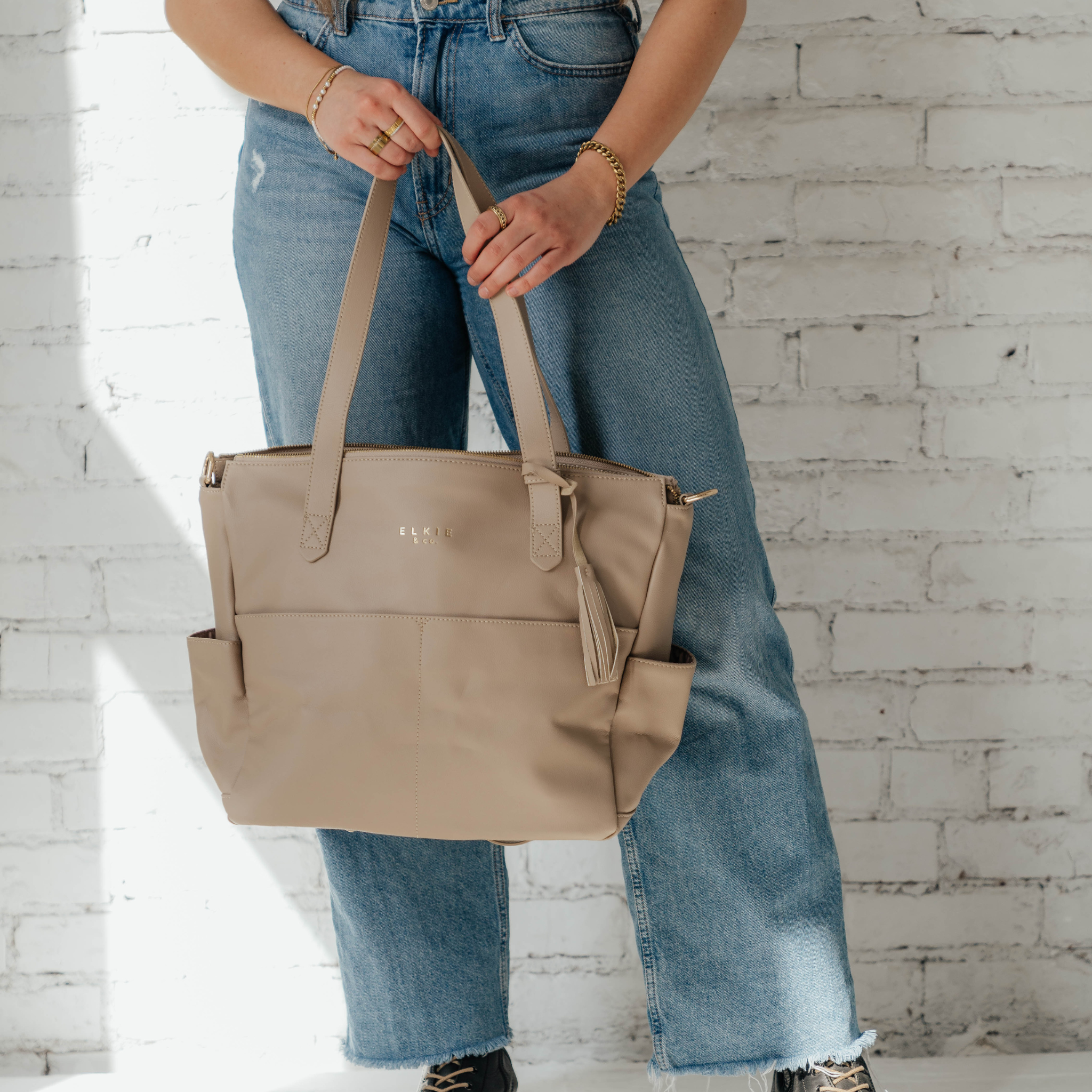 A person stands against a white brick wall, showcasing *The Aberdeen*, a stylish brown vegan leather tote with two front pockets and tassels.