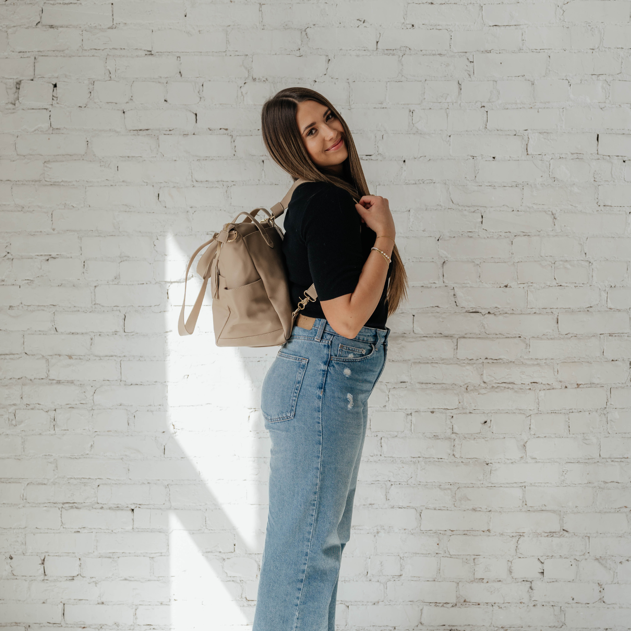 A person is standing against a white brick wall, showcasing The Aberdeen, a versatile brown leather backpack with multiple pockets.