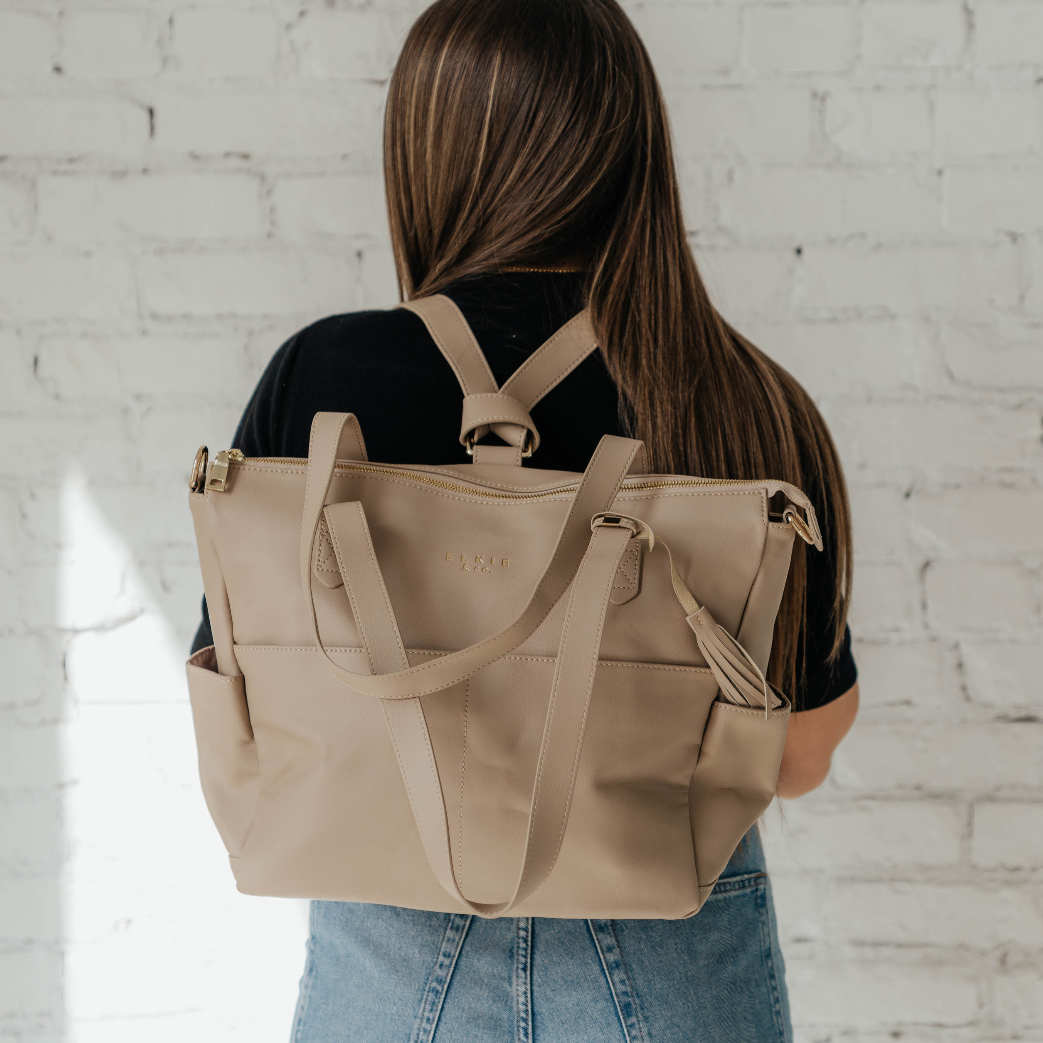 A person is standing against a white brick wall, showcasing The Aberdeen, a versatile brown leather backpack with multiple pockets.