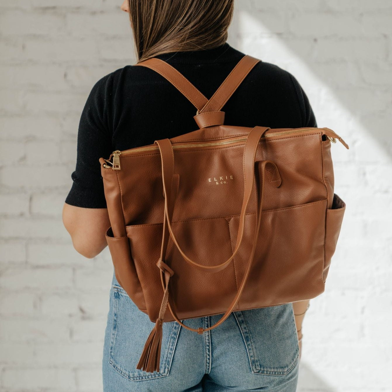 A person stands against a white brick wall, showcasing *The Aberdeen*, a stylish brown vegan leather tote with two front pockets and tassels.