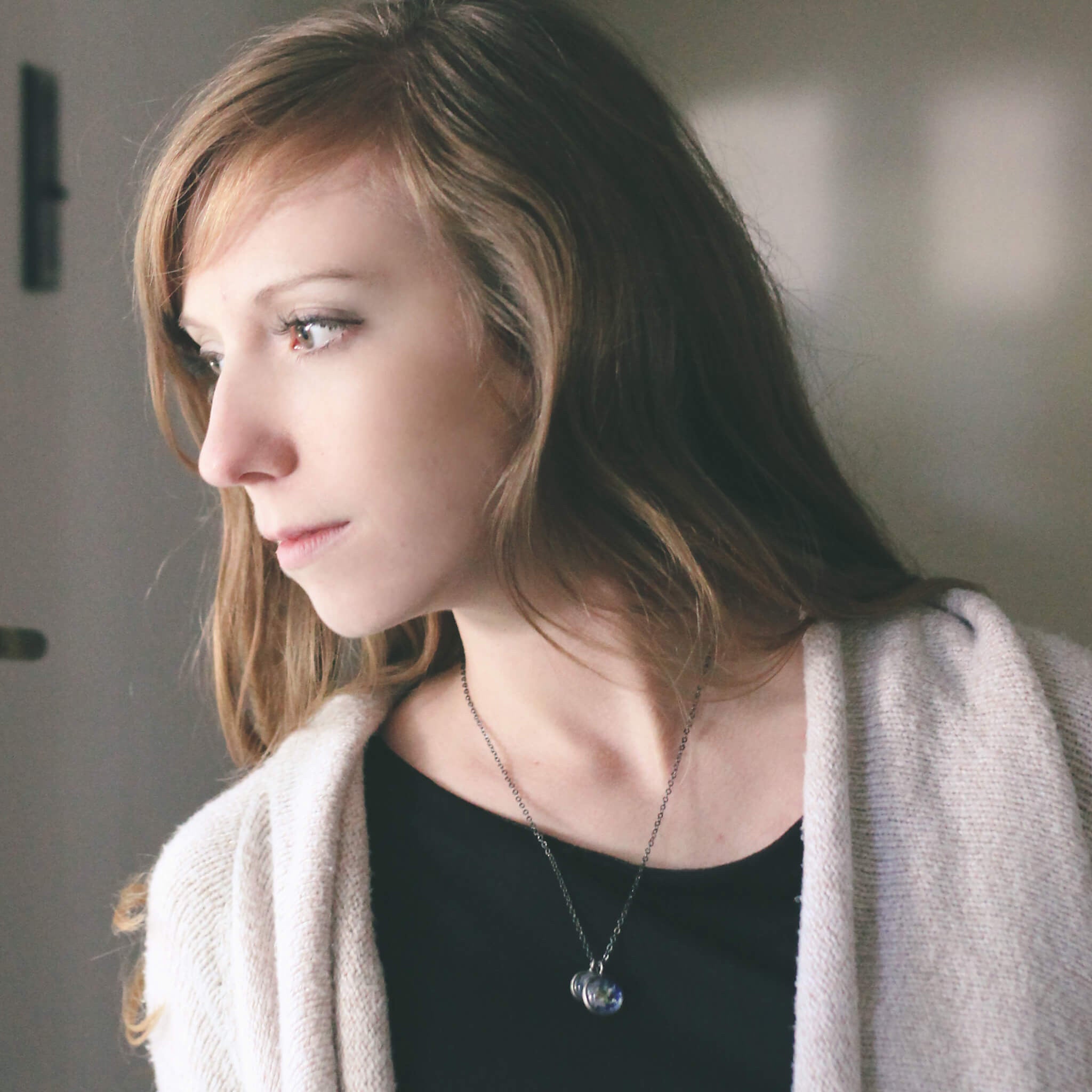 On a wooden surface, two silver Earth and Moon Layered Space Necklaces are adorned with pendants depicting Earth and the Moon. Encircled by small star-shaped embellishments, this celestial jewelry captures the ethereal beauty of our universe.